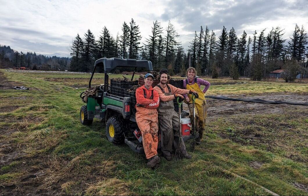 Three smiling people in dirty rain bibs with garden tools standing in front of an UTV in a farm field.