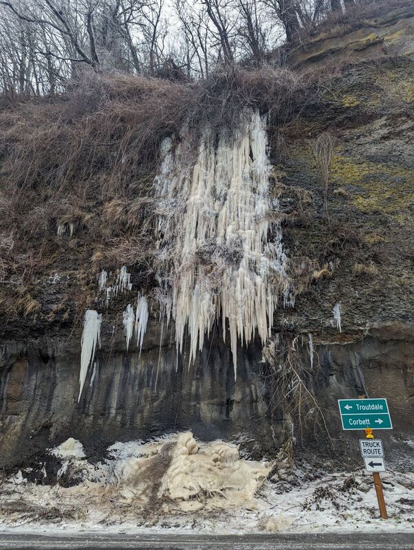 Picture of a rock cliff wall with icicles that look like a frozen waterfall. Street signs in front